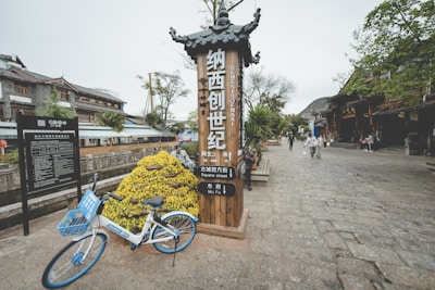 A cobblestone street in an old town with traditional architecture, featuring a wooden sign with Chinese characters and English translation. A parked blue and white bicycle is located near a mound of yellow flowers. Several people are strolling down the street lined with trees and wooden buildings.