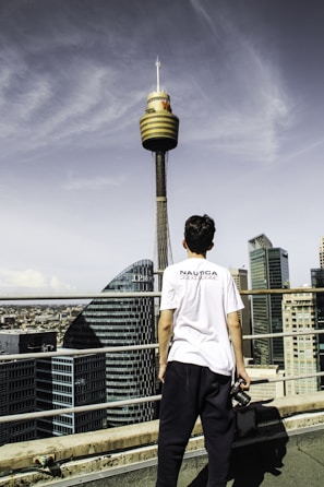 A person stands on a rooftop holding a camera, facing a city skyline dominated by a tall communication tower with a unique circular design. Modern skyscrapers surround the area under a partly cloudy sky.