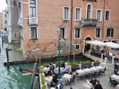 A picturesque canal-side scene with people dining at tables under umbrellas. The setting features a historic building with rustic walls and small balconies adorned with plants. A gondola is moored at the water's edge, adding a touch of classic Venetian charm. The water in the canal is a deep green color, and the area is lined with cobblestones and lampposts.
