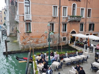 A picturesque canal-side scene with people dining at tables under umbrellas. The setting features a historic building with rustic walls and small balconies adorned with plants. A gondola is moored at the water's edge, adding a touch of classic Venetian charm. The water in the canal is a deep green color, and the area is lined with cobblestones and lampposts.