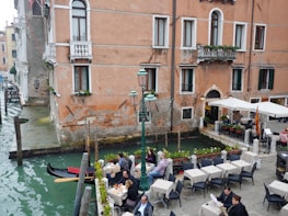 A picturesque canal-side scene with people dining at tables under umbrellas. The setting features a historic building with rustic walls and small balconies adorned with plants. A gondola is moored at the water's edge, adding a touch of classic Venetian charm. The water in the canal is a deep green color, and the area is lined with cobblestones and lampposts.