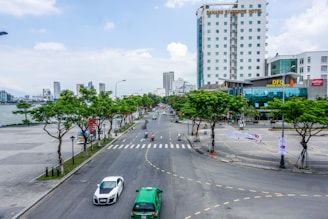 A wide urban road flanked by trees on both sides with a river visible to the left. There are several vehicles including cars and motorbikes on the road. On the right, a tall building with the name 'Danang Riverside Hotel' is prominent, alongside other modern structures including a complex labeled 'Danang Food Center'. The area is not very crowded with people, and the sky is partly cloudy.