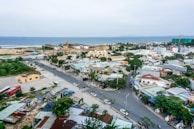 An aerial view of a town close to the ocean. The town has numerous buildings, including both residential and commercial structures. There are several roads with moving vehicles, and a few larger buildings with cranes suggesting ongoing construction. The background features a coastline with the sea meeting the horizon.