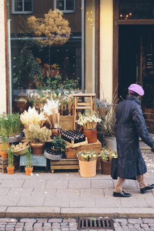 An outdoor display of a flower shop with various potted plants and dry flowers arranged on crates and tables. A woman in a long coat and purple beret walks past, holding a black bag. The shop's window reflects streetlights and greenery indoors.