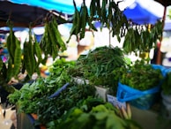 A market stall displaying a variety of fresh green vegetables and herbs. Long pods hang from the top, while bundles of leafy greens are stacked on the table below. The background features blurred market stalls under blue tents.