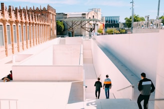A modern architectural setting features a geometric design with white walls and steps leading down to an unseen lower level. The area is bordered by a structure made of red brick with arched windows. Several people are walking or standing in the area, dressed casually. Trees and additional buildings are visible in the background under a clear blue sky.