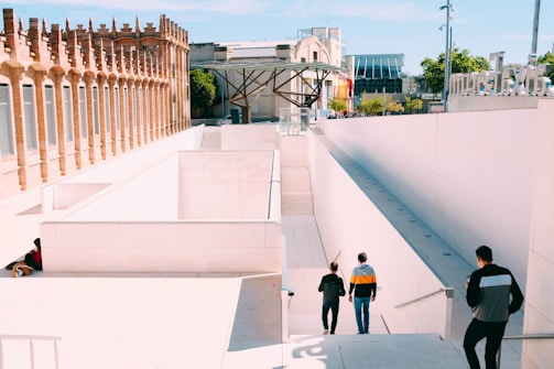 A modern architectural setting features a geometric design with white walls and steps leading down to an unseen lower level. The area is bordered by a structure made of red brick with arched windows. Several people are walking or standing in the area, dressed casually. Trees and additional buildings are visible in the background under a clear blue sky.