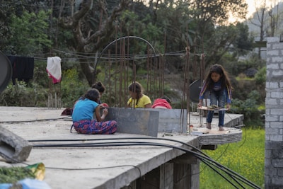 Four children are playing on an unfinished concrete structure, which appears to be a rooftop. One girl is standing with something in her hand, possibly a plate, while other children are seated, engaged in various activities. The background includes lush green trees and fields, indicating a rural setting. There are metal rods protruding from the concrete, suggesting ongoing construction. Pieces of cloth are hanging on wires, possibly for drying.