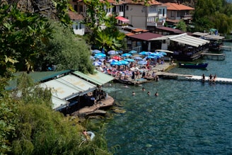 A picturesque lakeside view featuring a cluster of houses and small buildings with tiled roofs near the water. Numerous people are enjoying the sunny day on a waterside platform with colorful umbrellas and sun loungers. Several individuals are swimming in the clear blue lake. Trees and lush greenery form a natural boundary on one side, providing shade and a serene atmosphere.