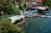 A picturesque lakeside view featuring a cluster of houses and small buildings with tiled roofs near the water. Numerous people are enjoying the sunny day on a waterside platform with colorful umbrellas and sun loungers. Several individuals are swimming in the clear blue lake. Trees and lush greenery form a natural boundary on one side, providing shade and a serene atmosphere.