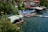 A picturesque lakeside view featuring a cluster of houses and small buildings with tiled roofs near the water. Numerous people are enjoying the sunny day on a waterside platform with colorful umbrellas and sun loungers. Several individuals are swimming in the clear blue lake. Trees and lush greenery form a natural boundary on one side, providing shade and a serene atmosphere.