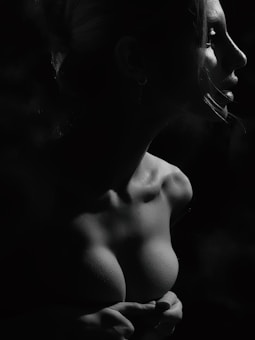 A dramatic black and white portrait of a woman in profile, highlighting her facial features and collarbone. The lighting casts deep shadows and accentuates the texture of her skin, creating a striking contrast.