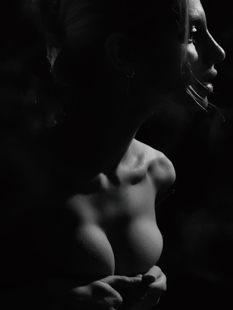A dramatic black and white portrait of a woman in profile, highlighting her facial features and collarbone. The lighting casts deep shadows and accentuates the texture of her skin, creating a striking contrast.