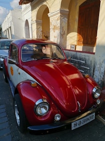 A classic red Volkswagen Beetle is parked in front of a weathered building with peeling paint and arched architecture. The car's glossy finish reflects the light, and there are a few other parked cars partially visible on the street.