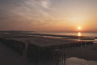 The image depicts a serene beach scene during sunset. The sun is low on the horizon, casting a warm, golden hue over the calm sea. The sky is partly cloudy with a soft gradient of colors from orange to light blue. Wooden posts are embedded in the sand, forming small barriers and pathways leading to the water. The landscape in the distance gently slopes down to meet the shoreline.