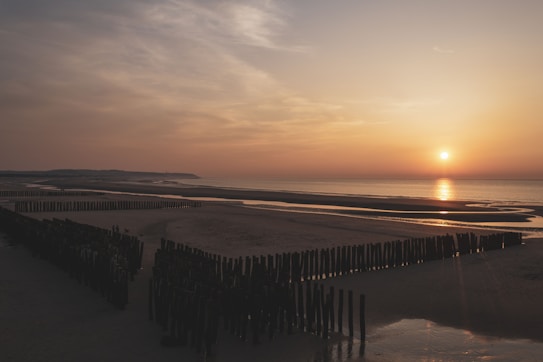 The image depicts a serene beach scene during sunset. The sun is low on the horizon, casting a warm, golden hue over the calm sea. The sky is partly cloudy with a soft gradient of colors from orange to light blue. Wooden posts are embedded in the sand, forming small barriers and pathways leading to the water. The landscape in the distance gently slopes down to meet the shoreline.
