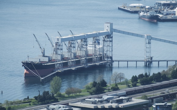 A large cargo ship docked at a port facility, equipped with multiple cranes for loading or unloading. The area is surrounded by a body of water, with several smaller boats and industrial structures in the distance. Lush greenery and trees border the water, with railway tracks visible in the foreground.