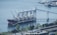 A large cargo ship docked at a port facility, equipped with multiple cranes for loading or unloading. The area is surrounded by a body of water, with several smaller boats and industrial structures in the distance. Lush greenery and trees border the water, with railway tracks visible in the foreground.