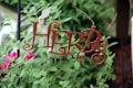 A decorative metal sign with the word 'HERBS' in curly letters is hanging among lush green plants. Bright pink flowers are visible in the background, adding a splash of color to the scene.