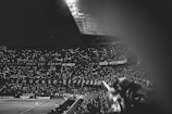 A crowded stadium filled with spectators, all of whom are holding up lights creating a sea of dots in the dark. The image is in black and white, giving it a nostalgic feel. There is a large banner displayed among the crowd, and a football match is taking place on the field below.
