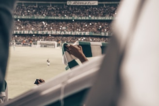 A crowded football stadium with fans holding a green and white scarf in focus. The field and players are visible in the background, with many audience members taking photos or cheering under the stadium lights.