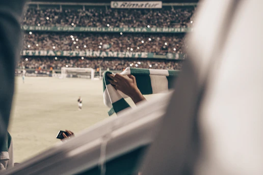 A vibrant football stadium under bright floodlights filled with cheering fans waving green and black scarves.