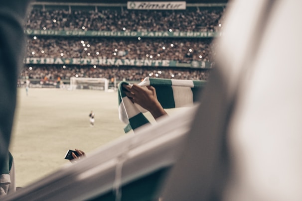 A crowded football stadium with fans holding a green and white scarf in focus. The field and players are visible in the background, with many audience members taking photos or cheering under the stadium lights.