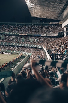 A large stadium filled with a crowd of enthusiastic spectators, many of whom are waving scarves and banners. The stadium is well-lit, with a section of the roof visible. There are security personnel in orange vests lined up along the sidelines near the field.