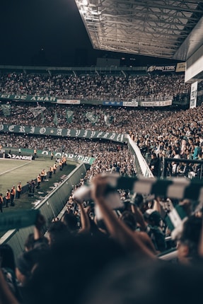 A large stadium filled with a crowd of enthusiastic spectators, many of whom are waving scarves and banners. The stadium is well-lit, with a section of the roof visible. There are security personnel in orange vests lined up along the sidelines near the field.