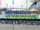 A large sign with the text '#DESTINATION UTTARAKHAND' stands prominently in an outdoor setting. The letters are bold and colorful, positioned against a backdrop of a stadium-like structure with flags of various colors around. People can be seen in the background walking across the area.