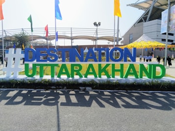 A large sign with the text '#DESTINATION UTTARAKHAND' stands prominently in an outdoor setting. The letters are bold and colorful, positioned against a backdrop of a stadium-like structure with flags of various colors around. People can be seen in the background walking across the area.