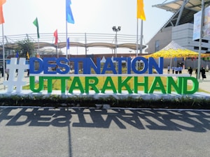 A large sign with the text '#DESTINATION UTTARAKHAND' stands prominently in an outdoor setting. The letters are bold and colorful, positioned against a backdrop of a stadium-like structure with flags of various colors around. People can be seen in the background walking across the area.
