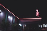 A dimly lit building features neon lights along the edges of its roof with a neon sign shaped like a bowling pin displaying the word 'BOWL'. Trees and plants are visible in the foreground under bright exterior lights.