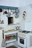 A small kitchen with a gas stove, various pots and pans hanging on hooks, and white tiled walls. There's a countertop with scattered kitchen items including a pot, dishes, and a lemon. A red bucket and a metal bowl are on the lower shelf. The refrigerator door has towels draped over and a kitchen towel hangs from the counter. Oven mitts and decorative pot holders are on the wall.