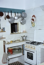 A small kitchen with a gas stove, various pots and pans hanging on hooks, and white tiled walls. There's a countertop with scattered kitchen items including a pot, dishes, and a lemon. A red bucket and a metal bowl are on the lower shelf. The refrigerator door has towels draped over and a kitchen towel hangs from the counter. Oven mitts and decorative pot holders are on the wall.