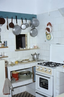 A small kitchen with a gas stove, various pots and pans hanging on hooks, and white tiled walls. There's a countertop with scattered kitchen items including a pot, dishes, and a lemon. A red bucket and a metal bowl are on the lower shelf. The refrigerator door has towels draped over and a kitchen towel hangs from the counter. Oven mitts and decorative pot holders are on the wall.