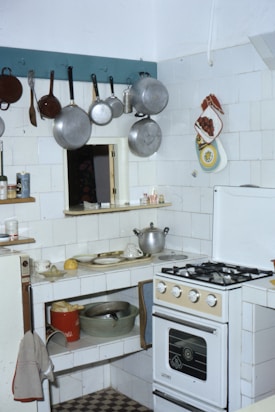 A small kitchen with a gas stove, various pots and pans hanging on hooks, and white tiled walls. There's a countertop with scattered kitchen items including a pot, dishes, and a lemon. A red bucket and a metal bowl are on the lower shelf. The refrigerator door has towels draped over and a kitchen towel hangs from the counter. Oven mitts and decorative pot holders are on the wall.
