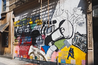 A vibrant mural painted on the exterior wall of a building. The mural features bold graphics and text related to social justice and civil rights. It includes an image of a person using a large video camera, surrounded by various text excerpts about rights and justice. The words 'KNOW YOUR RIGHTS' are prominently displayed on the mural. The artwork is a mix of colorful geometric shapes and monochrome drawings of people.