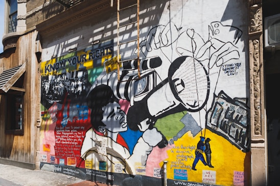 A vibrant mural painted on the exterior wall of a building. The mural features bold graphics and text related to social justice and civil rights. It includes an image of a person using a large video camera, surrounded by various text excerpts about rights and justice. The words 'KNOW YOUR RIGHTS' are prominently displayed on the mural. The artwork is a mix of colorful geometric shapes and monochrome drawings of people.
