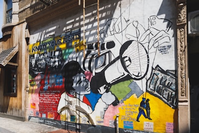A vibrant mural painted on the exterior wall of a building. The mural features bold graphics and text related to social justice and civil rights. It includes an image of a person using a large video camera, surrounded by various text excerpts about rights and justice. The words 'KNOW YOUR RIGHTS' are prominently displayed on the mural. The artwork is a mix of colorful geometric shapes and monochrome drawings of people.
