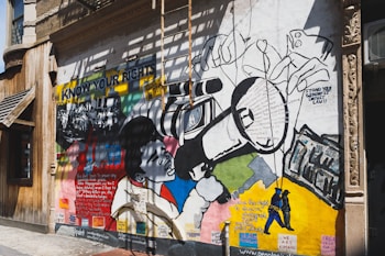 A vibrant mural painted on the exterior wall of a building. The mural features bold graphics and text related to social justice and civil rights. It includes an image of a person using a large video camera, surrounded by various text excerpts about rights and justice. The words 'KNOW YOUR RIGHTS' are prominently displayed on the mural. The artwork is a mix of colorful geometric shapes and monochrome drawings of people.