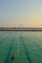 A swimming pool with lanes is filled with several people swimming. The water has a greenish hue, and the sky is clear with a subtle gradient from blue to orange at the horizon, suggesting either sunrise or sunset. In the background, there is a body of water and a distant view of a bridge or pier with structures that appear industrial.