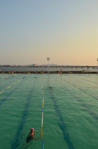 A swimming pool with lanes is filled with several people swimming. The water has a greenish hue, and the sky is clear with a subtle gradient from blue to orange at the horizon, suggesting either sunrise or sunset. In the background, there is a body of water and a distant view of a bridge or pier with structures that appear industrial.