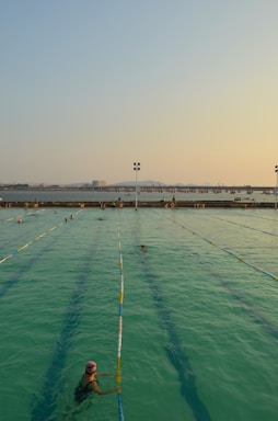A swimming pool with lanes is filled with several people swimming. The water has a greenish hue, and the sky is clear with a subtle gradient from blue to orange at the horizon, suggesting either sunrise or sunset. In the background, there is a body of water and a distant view of a bridge or pier with structures that appear industrial.
