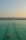 A swimming pool with lanes is filled with several people swimming. The water has a greenish hue, and the sky is clear with a subtle gradient from blue to orange at the horizon, suggesting either sunrise or sunset. In the background, there is a body of water and a distant view of a bridge or pier with structures that appear industrial.