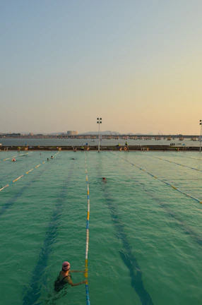 A swimming pool with lanes is filled with several people swimming. The water has a greenish hue, and the sky is clear with a subtle gradient from blue to orange at the horizon, suggesting either sunrise or sunset. In the background, there is a body of water and a distant view of a bridge or pier with structures that appear industrial.