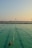 A swimming pool with lanes is filled with several people swimming. The water has a greenish hue, and the sky is clear with a subtle gradient from blue to orange at the horizon, suggesting either sunrise or sunset. In the background, there is a body of water and a distant view of a bridge or pier with structures that appear industrial.