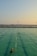 A swimming pool with lanes is filled with several people swimming. The water has a greenish hue, and the sky is clear with a subtle gradient from blue to orange at the horizon, suggesting either sunrise or sunset. In the background, there is a body of water and a distant view of a bridge or pier with structures that appear industrial.