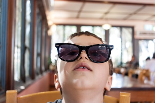 A young child wearing oversized sunglasses sits in a wooden chair, gazing upwards. The background features a blurred view of a dining area with large windows and natural light filtering in. The scene appears to take place indoors in a casual setting.