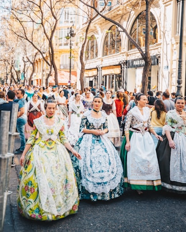 A lively street scene during a cultural festival where women are dressed in elaborate traditional dresses. The intricate patterns and vivid colors of the attire stand out. A diverse group of people gathers in the background amidst a historic cityscape with ornate buildings and leafless trees.